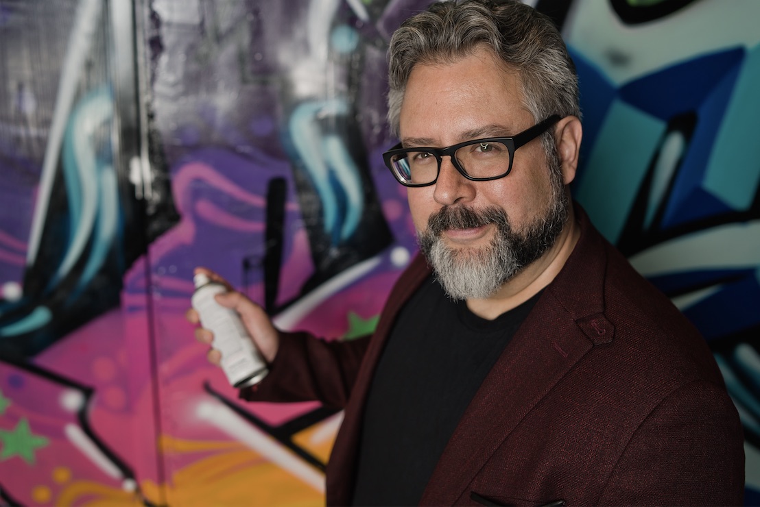 Man in glasses holding spray paint can in front of wall of graffiti